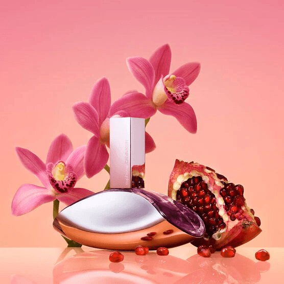 Cena Euphoria: frasco prata-violeta cercado de orquídeas rosas, romã aberta com sementes rubi sobre superfície coral