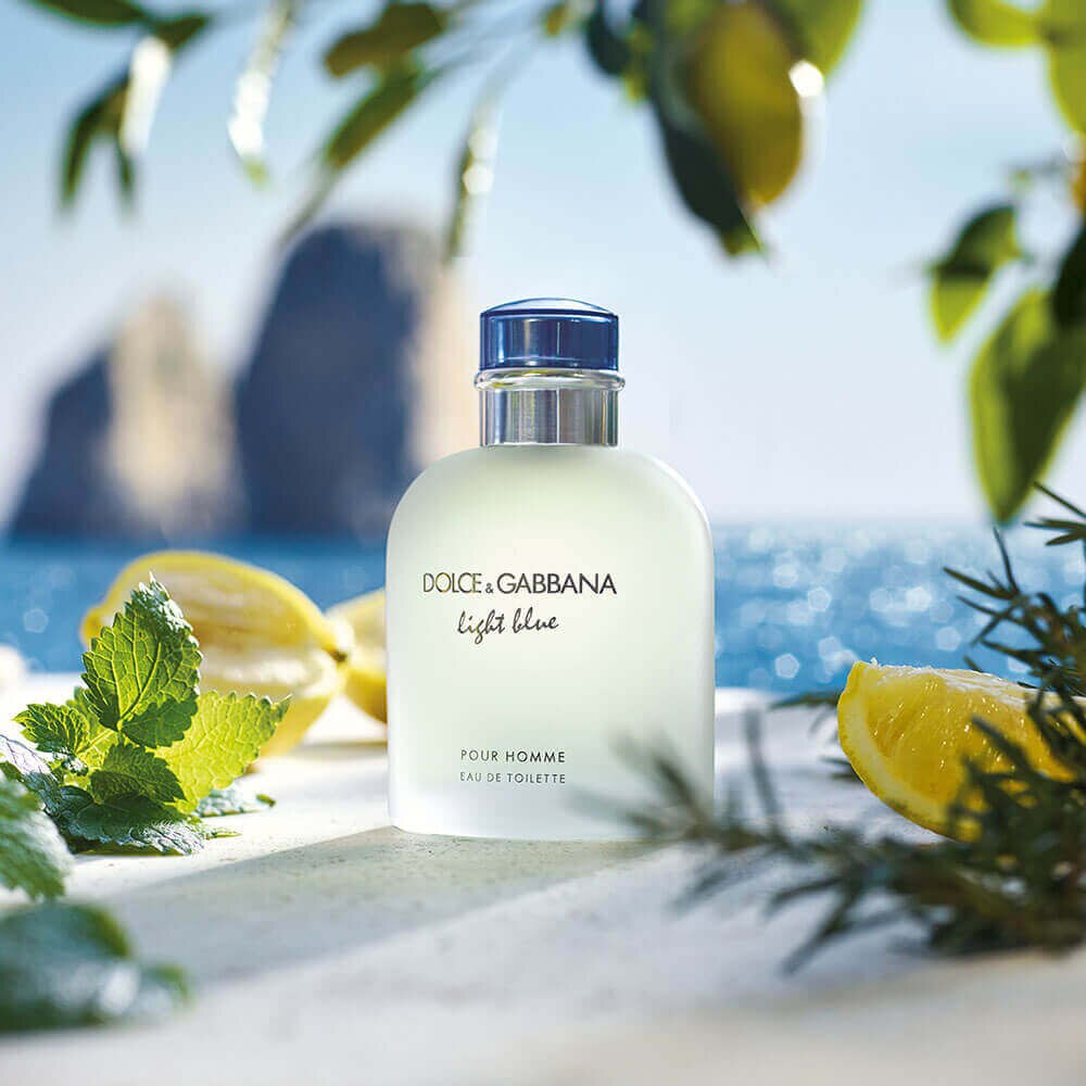 Cena oficial Light Blue: o frasco frosted com tampa azul sobre pedra branca de Capri, com folhas de limoeiro, fatias de limão siciliano, alecrim e os Faraglioni ao fundo na água azul mediterrânea