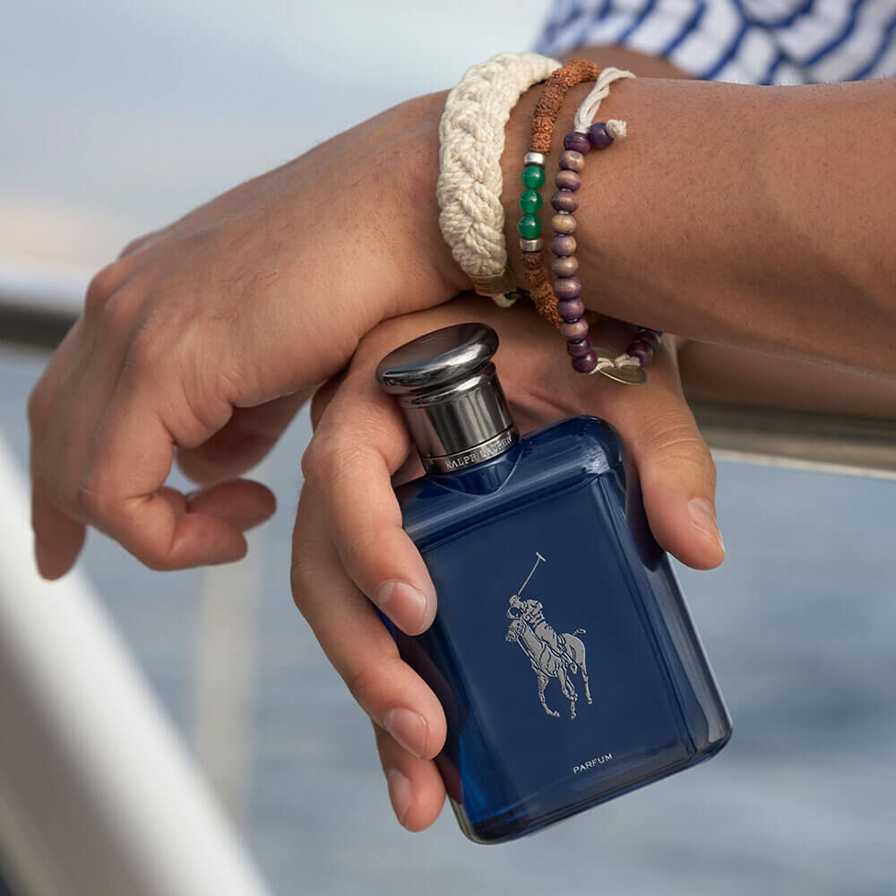 Polo Blue Parfum nas mãos de um homem sobre o convés de um barco à vela, com a água ao fundo no fim da tarde