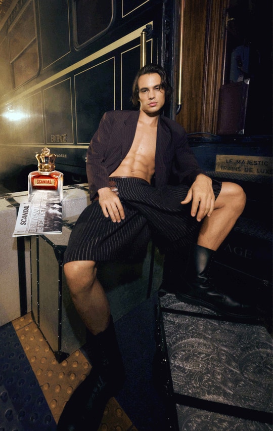 Modelo da campanha Scandal pour Homme Elixir sentado nos degraus do vagão Le Majestic, em alfaiataria listrada preta, com o frasco coroado de Scandal Elixir ao lado.